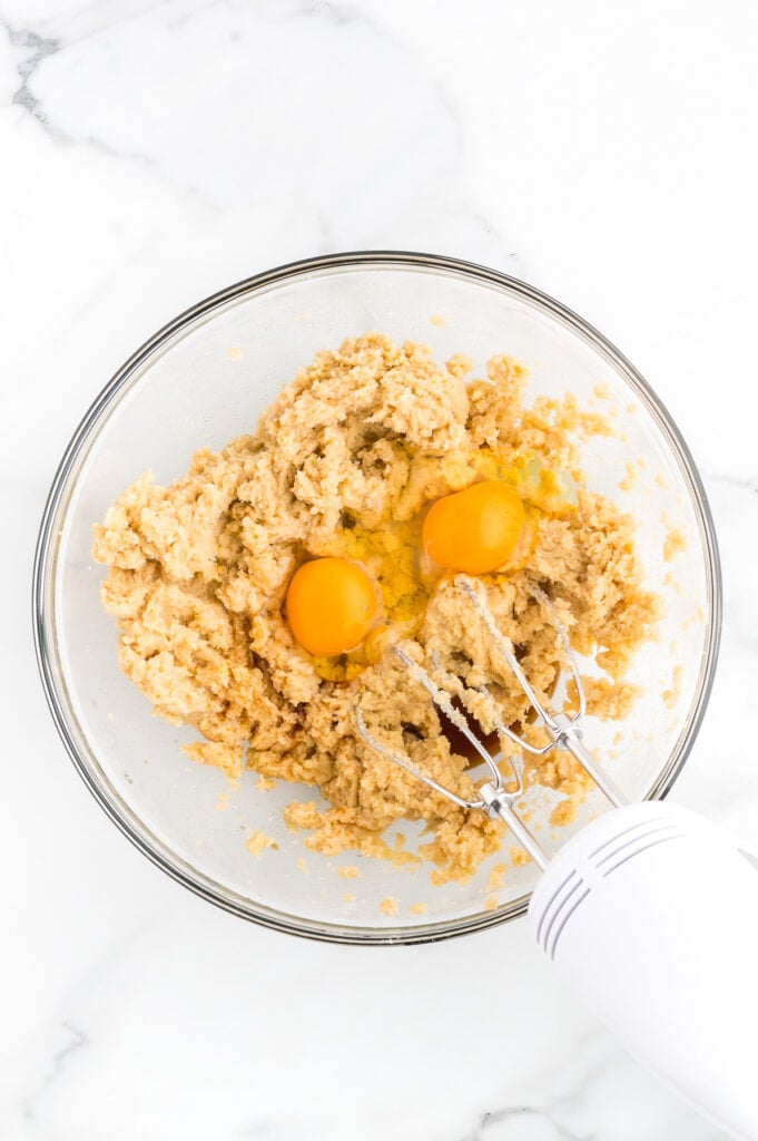 A glass bowl with cookie dough mixture, two whole eggs, and a hand mixer on a white marble surface.