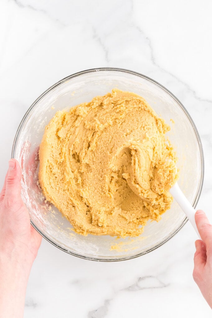 A person stirring thick cookie dough in a clear glass bowl with a white spatula on a white marble surface.