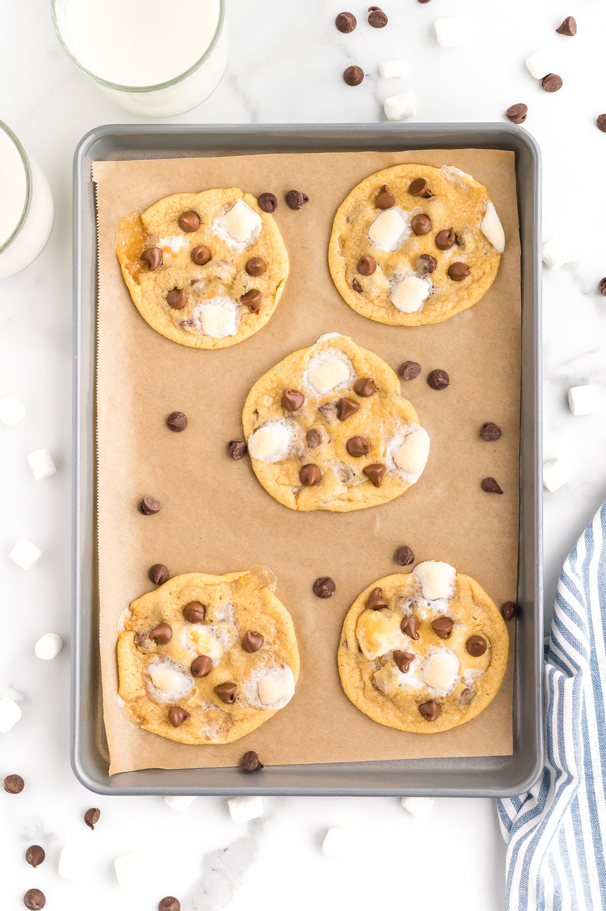 A baking tray lined with parchment paper holds five cookies topped with marshmallows and chocolate chips; extra chocolate chips and marshmallows are scattered around.