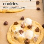 A chocolate chip cookie with melted marshmallows and chocolate chips on parchment paper, next to a baking tray.