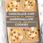 A baking sheet with six chocolate chip marshmallow cookies on parchment paper, surrounded by mini marshmallows and chocolate chips.