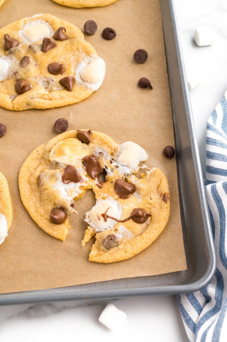 A baking sheet with chocolate chip cookies topped with marshmallows, one cookie broken in half, on parchment paper next to a blue and white striped cloth.