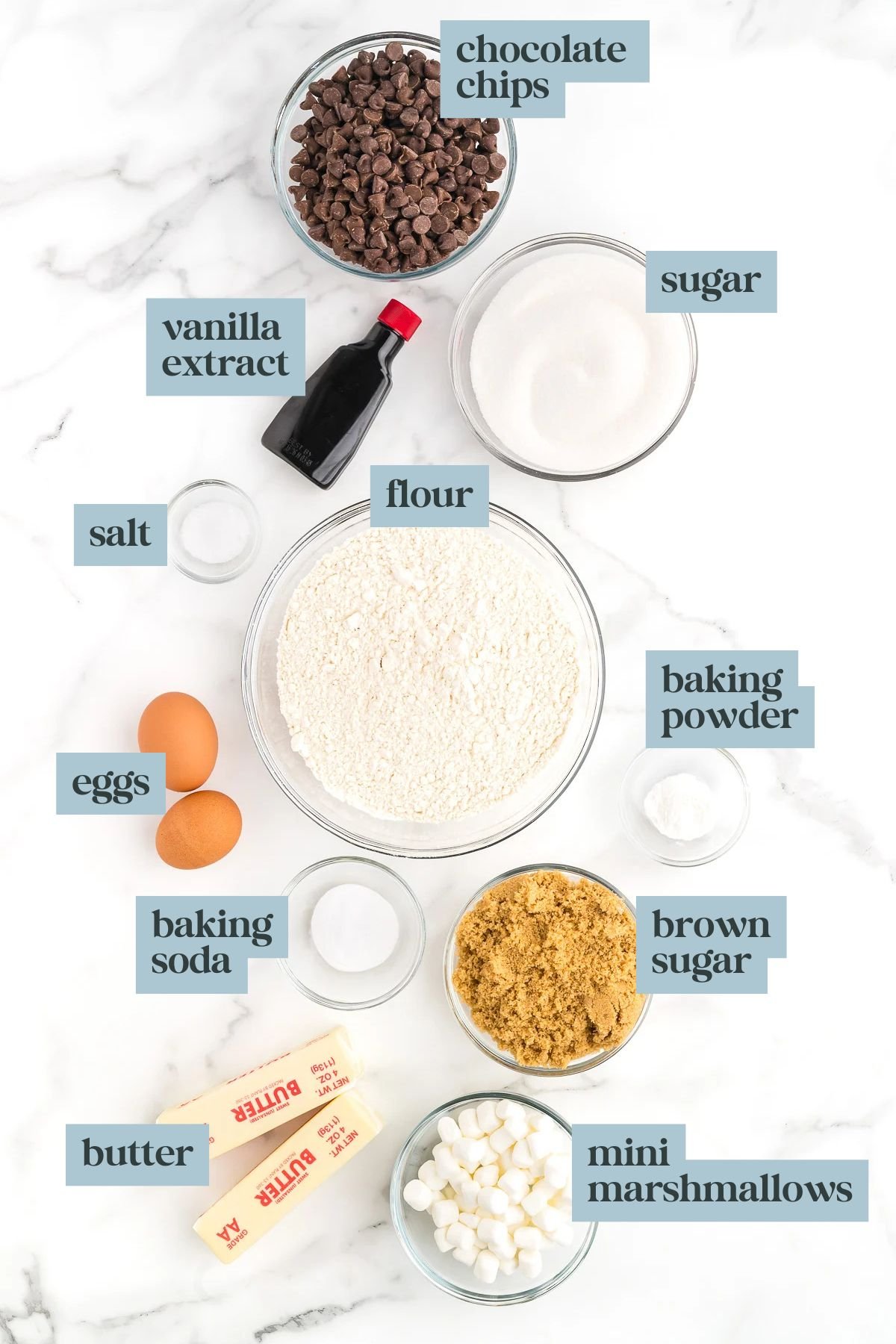 Baking ingredients arranged on a white surface, including chocolate chips, flour, sugar, brown sugar, mini marshmallows, eggs, butter, baking soda, baking powder, salt, and vanilla extract.