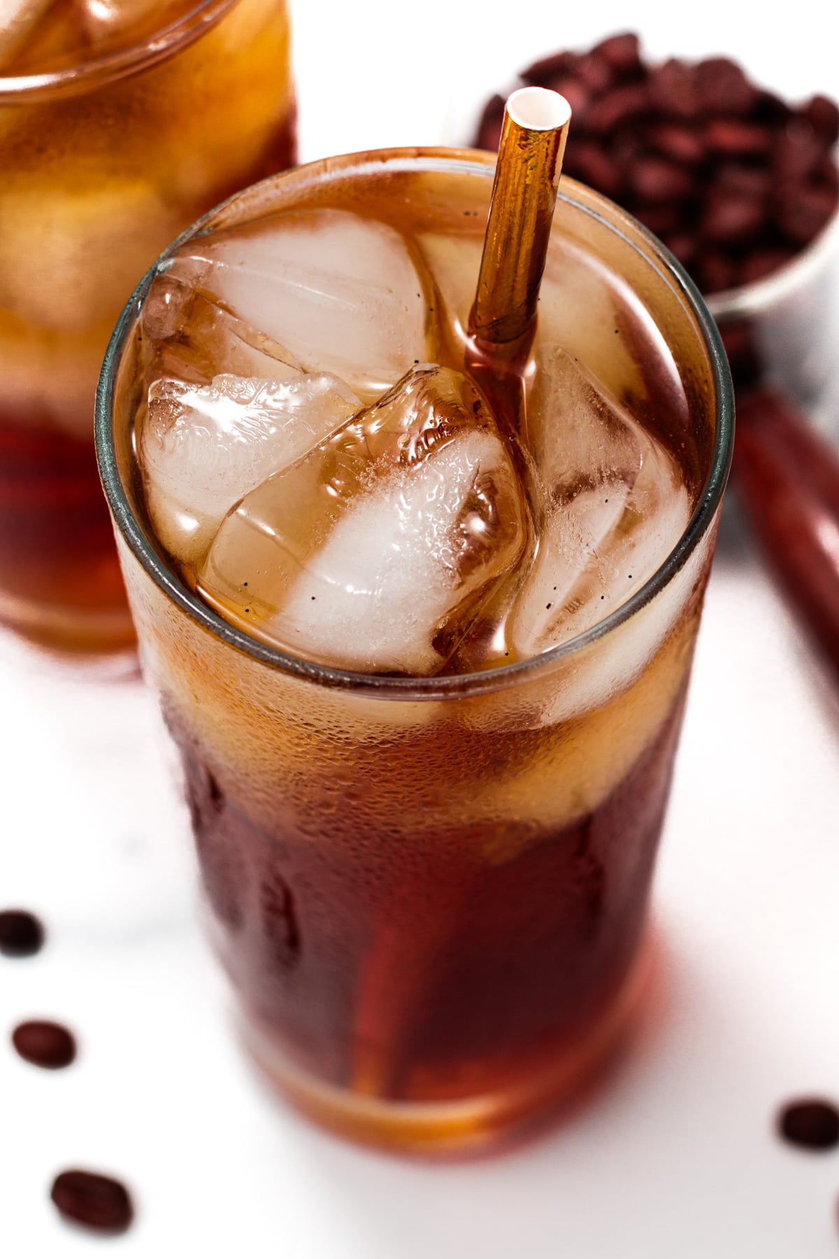 A glass of iced coffee with a brown straw, filled with ice cubes. Coffee beans are scattered nearby and a small bowl of beans is visible in the background.