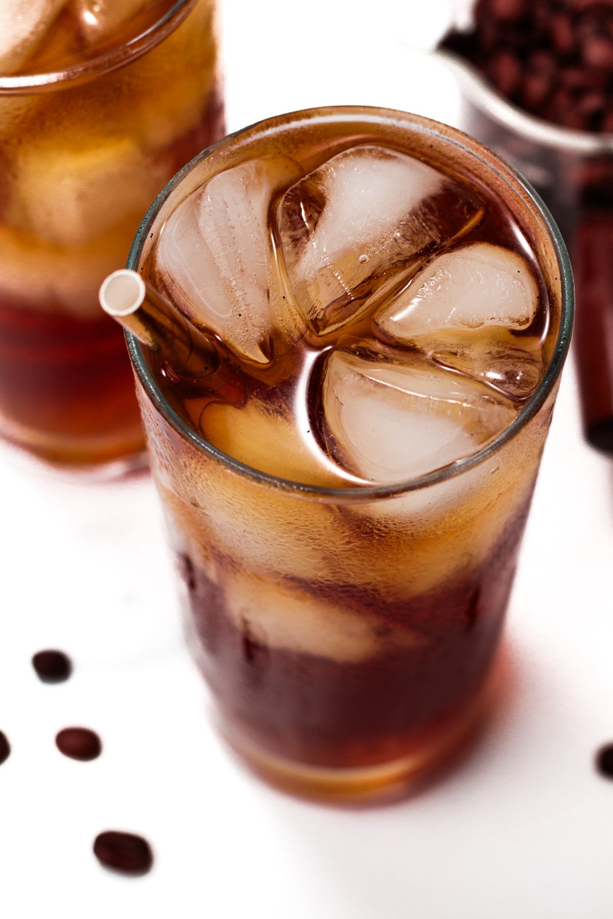 A glass of iced coffee with a straw and ice cubes, with another glass and coffee beans in the background.