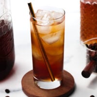 A glass of iced coffee with a straw sits on a wooden coaster, surrounded by a jar of coffee, a carafe, and a small container of coffee beans.