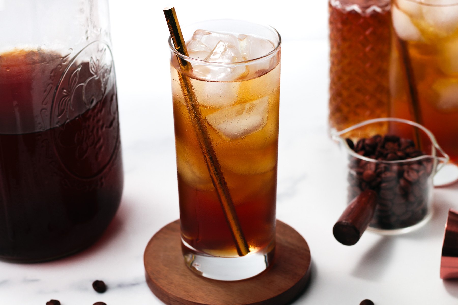 A glass of iced coffee with a straw sits on a wooden coaster, surrounded by a jar of coffee, a carafe, and a small container of coffee beans.