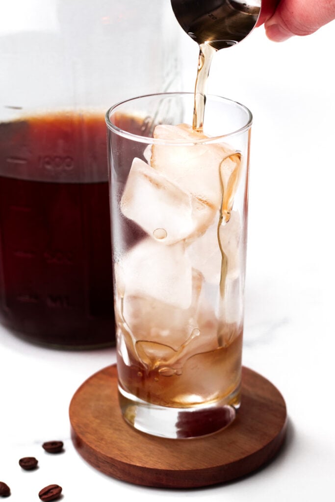A hand pours liquid into a tall glass filled with ice cubes. A large container with more liquid and a few coffee beans are visible nearby.