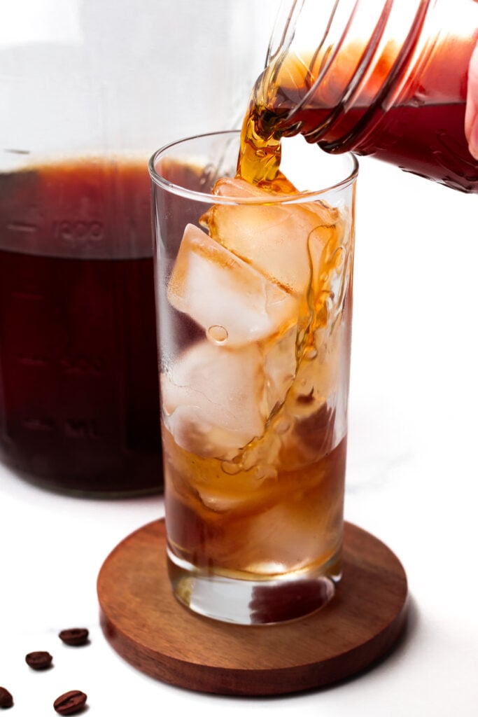 Cold brew coffee is being poured from a jar into a glass filled with ice cubes, with coffee beans and a carafe in the background.