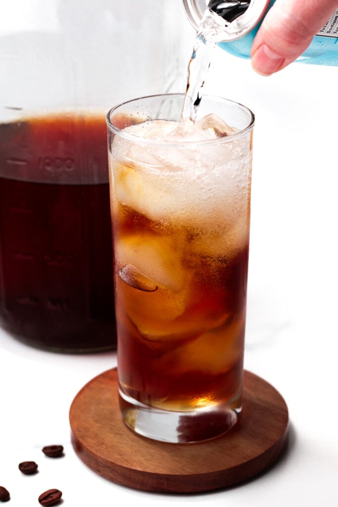 A hand pours a clear carbonated liquid into a tall glass of iced coffee on a wooden coaster, with a jar of coffee and coffee beans nearby.