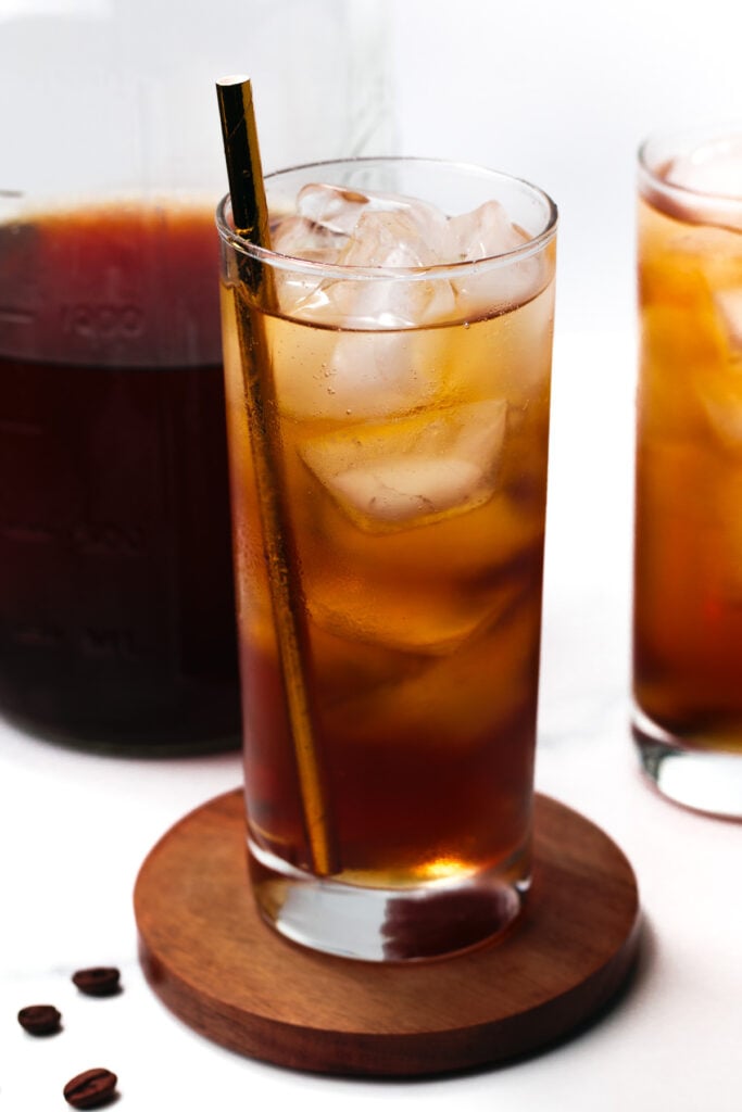 A tall glass of iced coffee with a metal straw on a wooden coaster, surrounded by a glass pitcher and another glass, with a few coffee beans nearby.