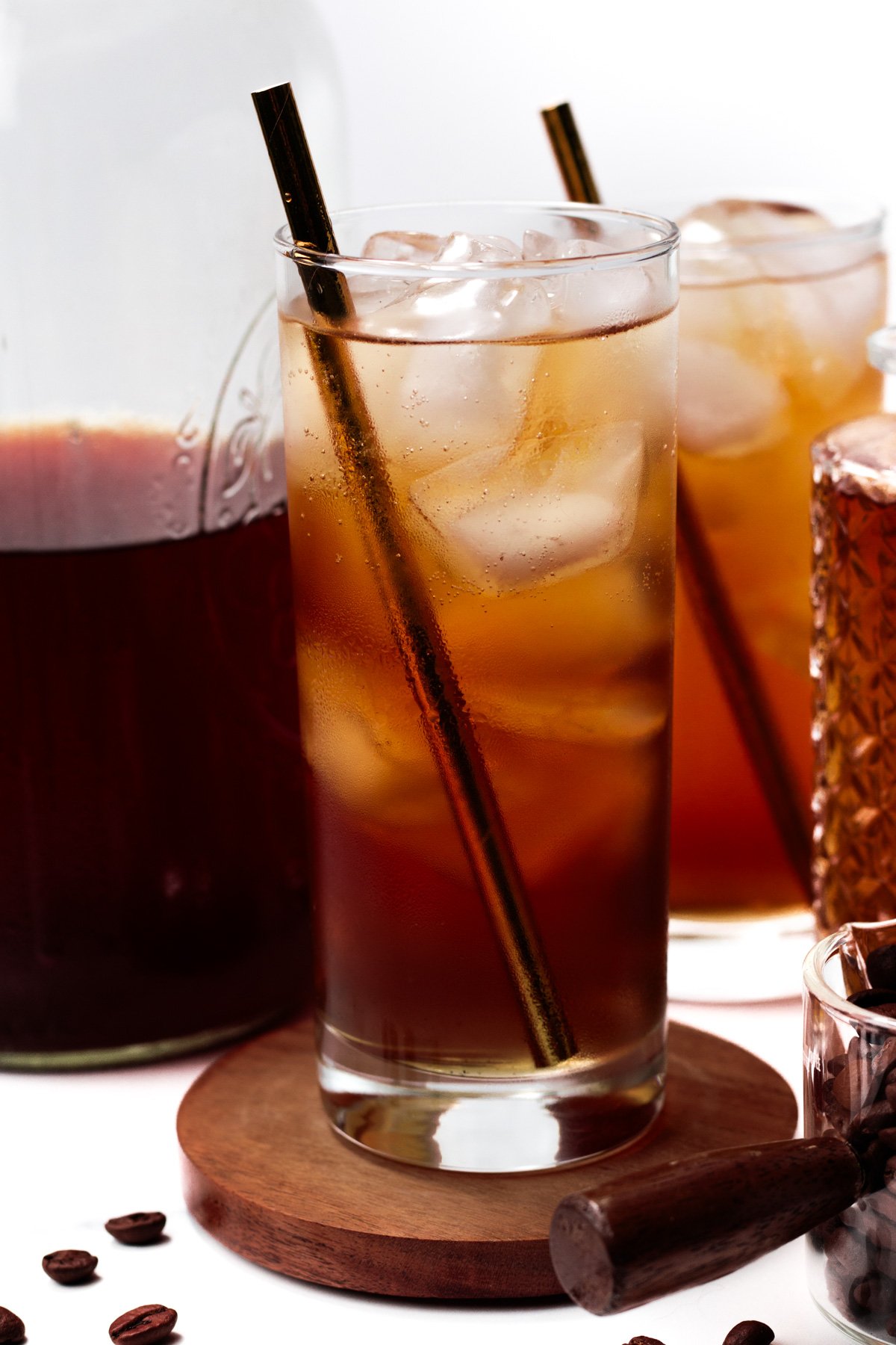 A glass of iced coffee with a metal straw sits on a wooden coaster, surrounded by a jar, another glass, coffee beans, and a small jar on a white background.