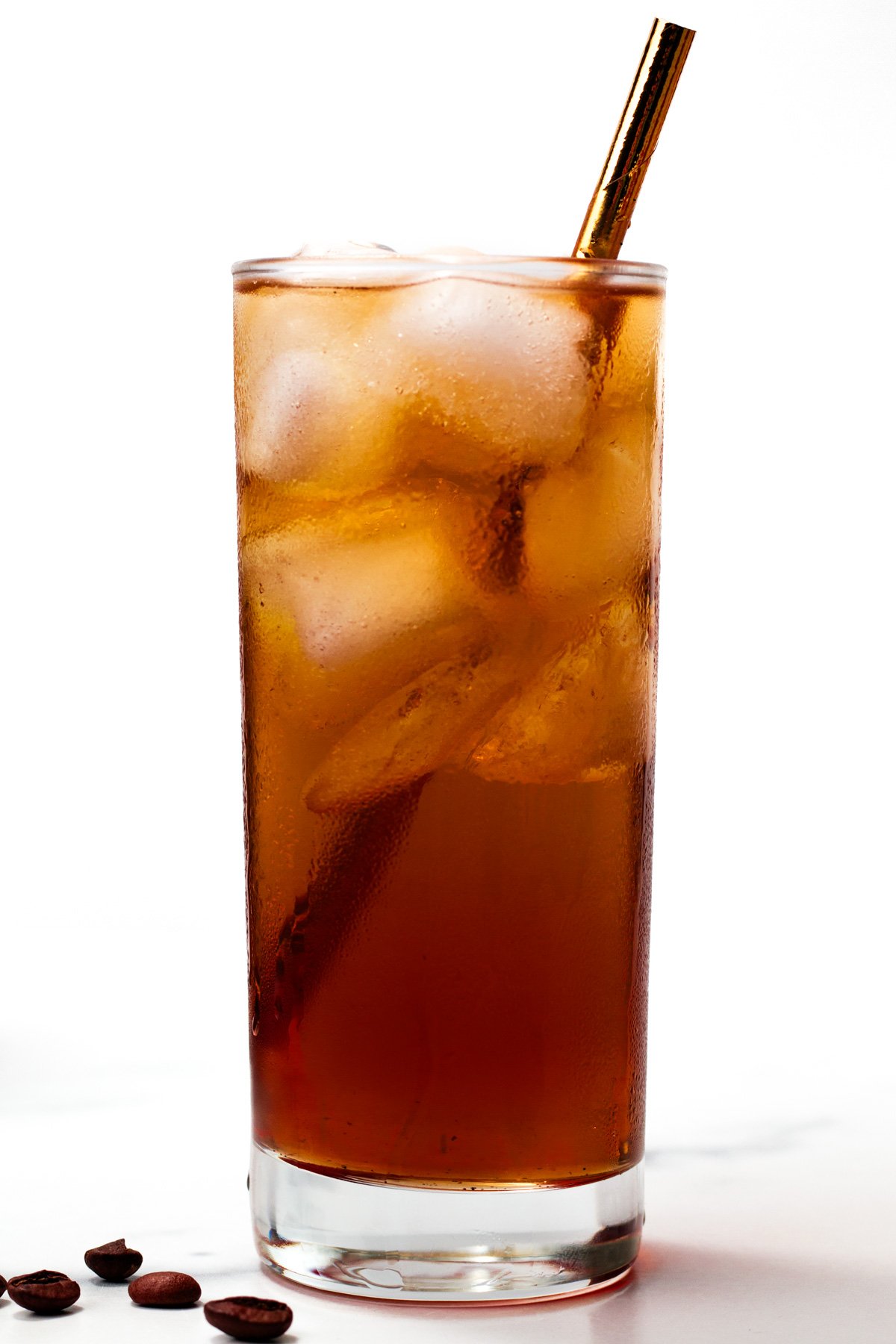 A tall glass filled with iced coffee and a metal straw, with a few coffee beans placed on the surface nearby.