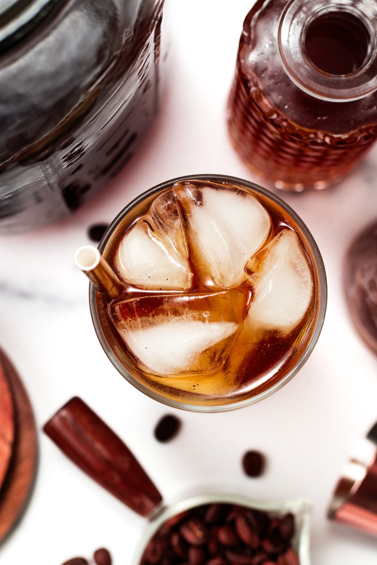A glass of iced coffee with a straw, surrounded by a jar, a bottle, coffee beans, and a small pitcher on a white surface.