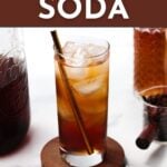 A glass of refreshing coffee soda with a straw sits on a round coaster, surrounded by glass jars and coffee beans. Text reads “COFFEE SODA—GET THE RECIPE—feastandwest.com.”.