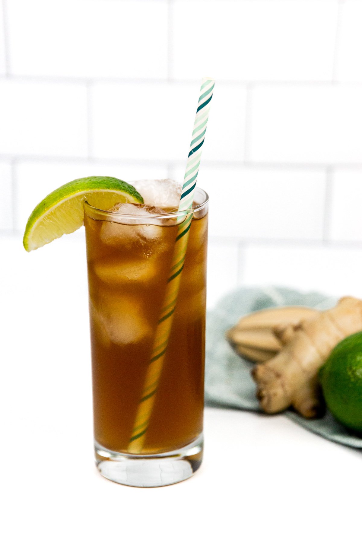 A tall glass of iced brown beverage with a lime wedge and a striped straw, placed next to fresh ginger and a lime on a white surface.