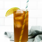 A tall glass of iced tea with a striped straw, ice cubes, and a lime wedge garnish, placed in front of a white tile background and a folded gray cloth.