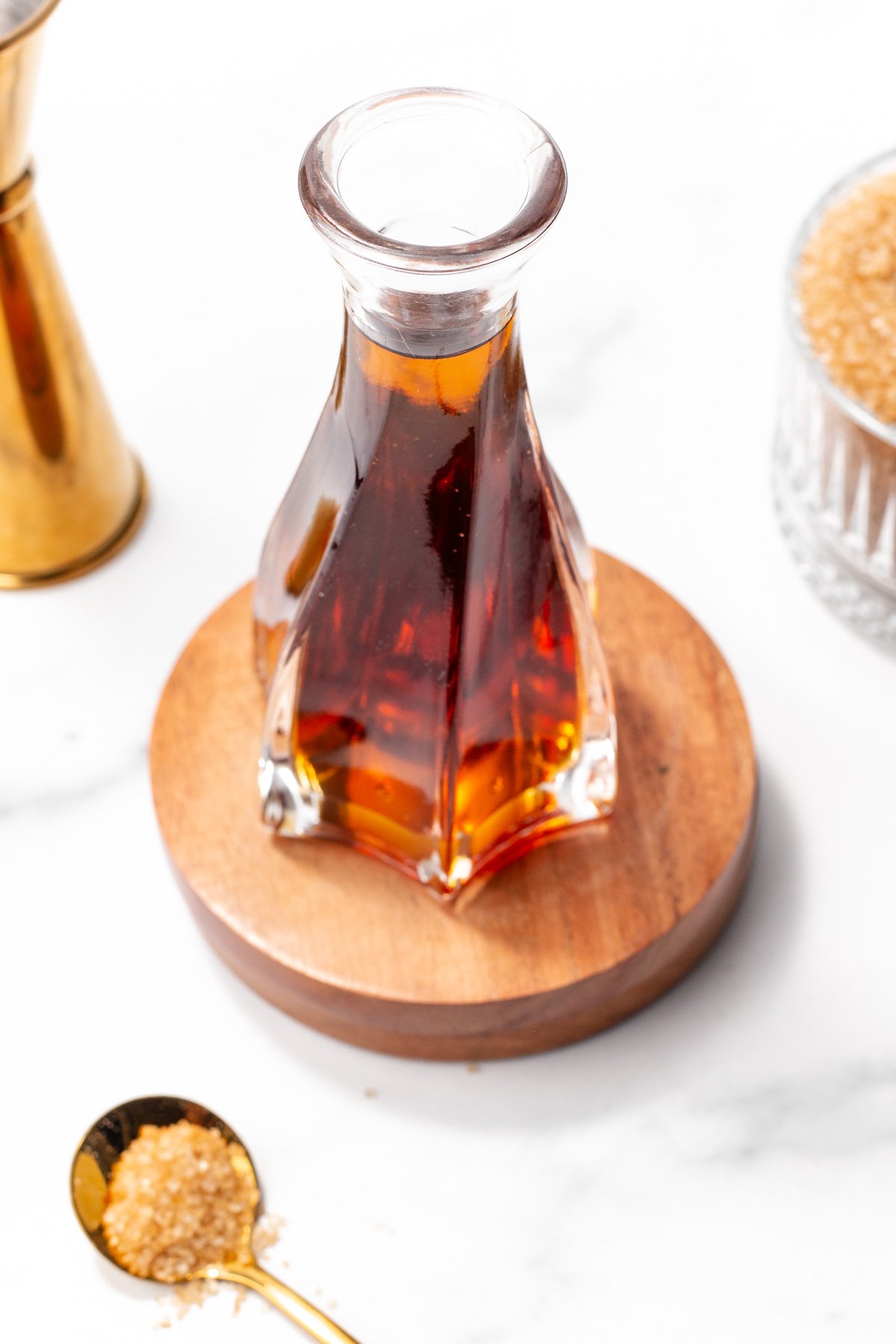 A glass carafe filled with amber liquid sits on a wooden coaster, surrounded by a spoonful and a bowl of brown sugar, and a brass container on a white surface.