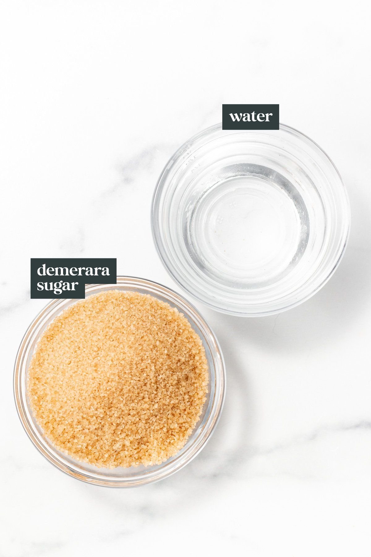 Two glass bowls on a white surface: one containing water and the other filled with demerara sugar. Each bowl is labeled accordingly.