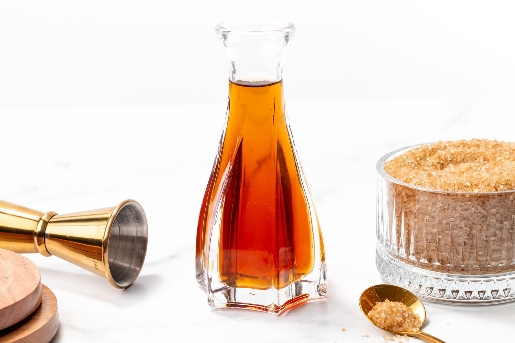 A glass decanter filled with amber liquid sits between a gold jigger and a bowl of brown sugar on a white surface.