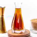 A glass decanter filled with amber liquid is placed on a wooden base, surrounded by a jigger, a glass bowl of brown sugar, and a gold spoon with brown sugar.