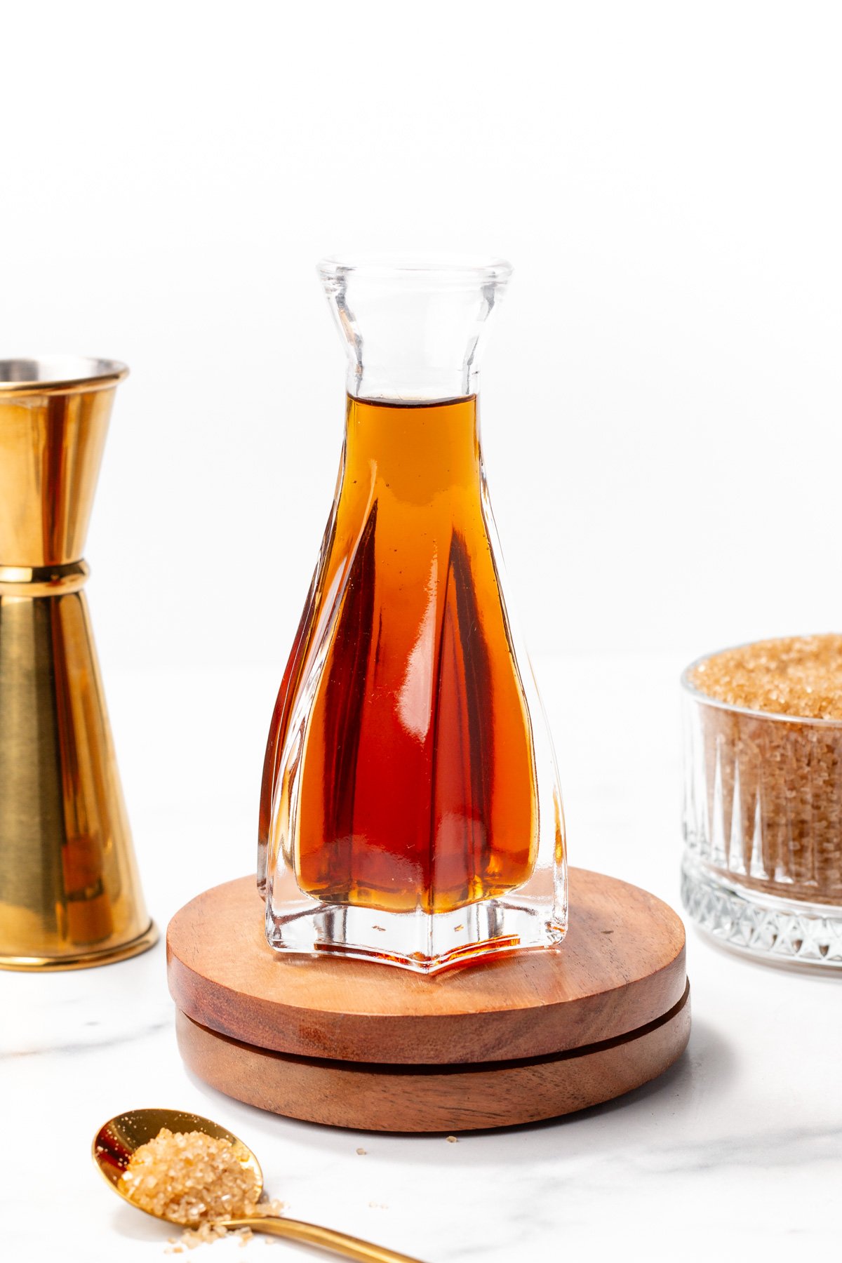 A glass decanter filled with amber liquid is placed on a wooden base, surrounded by a jigger, a glass bowl of brown sugar, and a gold spoon with brown sugar.