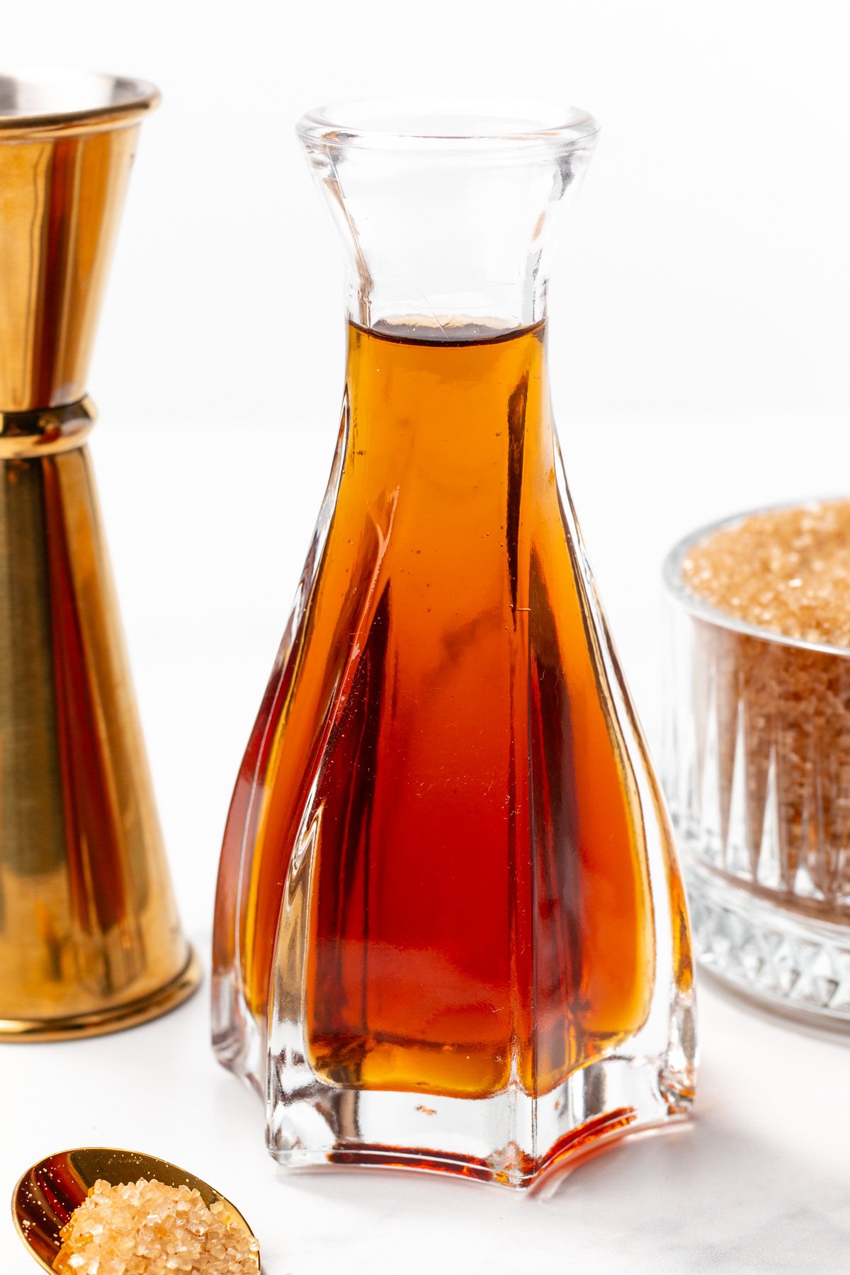 A clear glass decanter filled with amber-colored liquid is placed next to a metal jigger, a bowl of brown sugar, and a spoon containing brown sugar.
