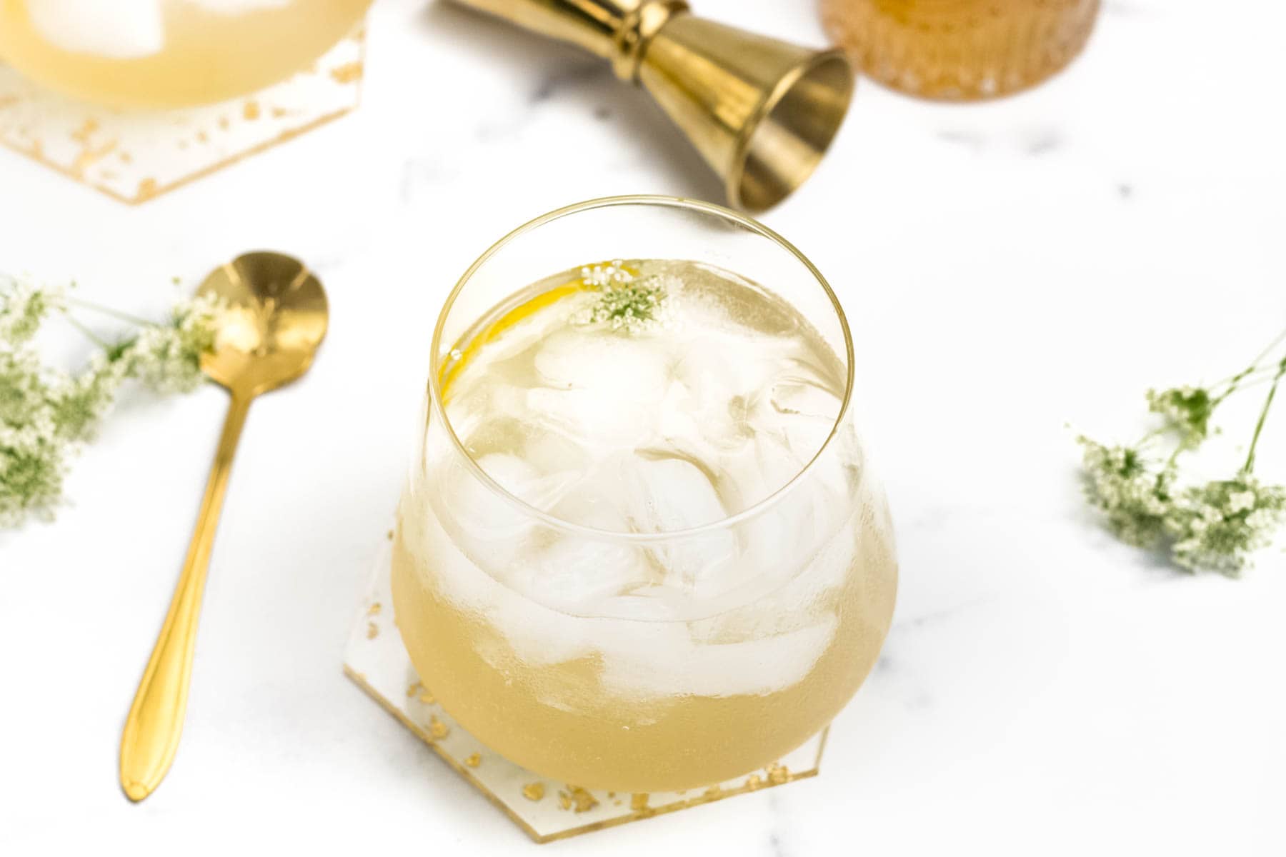 A glass of light-colored cocktail with ice sits on a coaster, next to a gold spoon and a gold jigger on a white surface with small white flowers.
