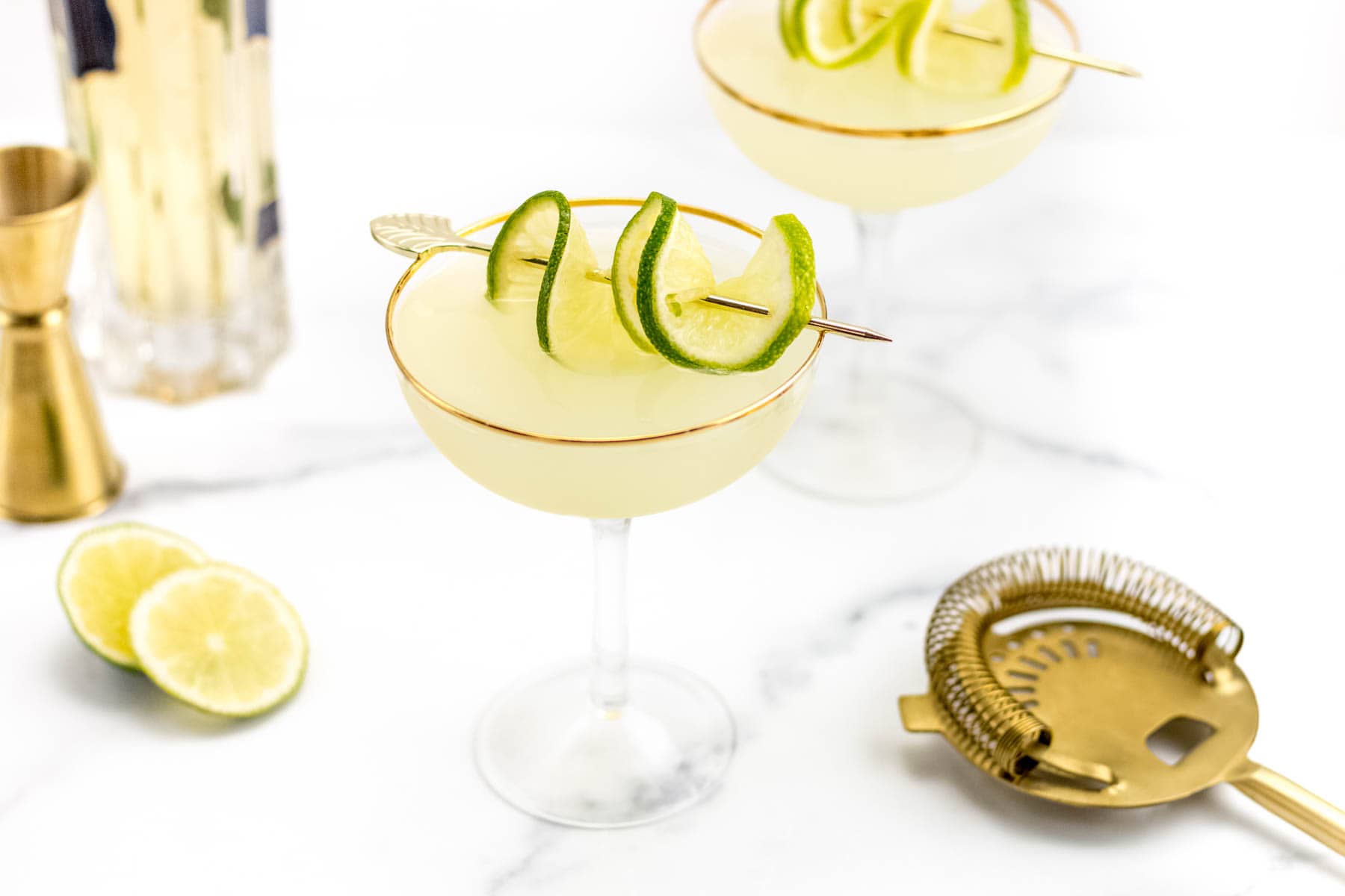 A coupe glass filled with a pale yellow cocktail, garnished with lime slices on a skewer. Gold bar tools and lime halves are nearby on a white marble surface.
