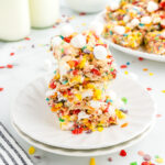 Three stacked Fruity Pebbles cereal treats with mini marshmallows on a white plate, with milk bottles and more treats in the background.