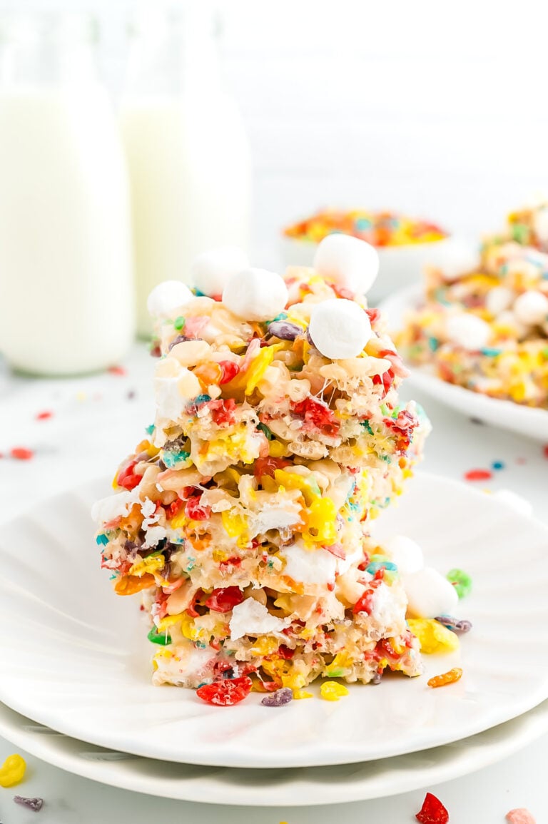 Three colorful cereal marshmallow bars stacked on a white plate, with mini marshmallows on top and two bottles of milk in the background.
