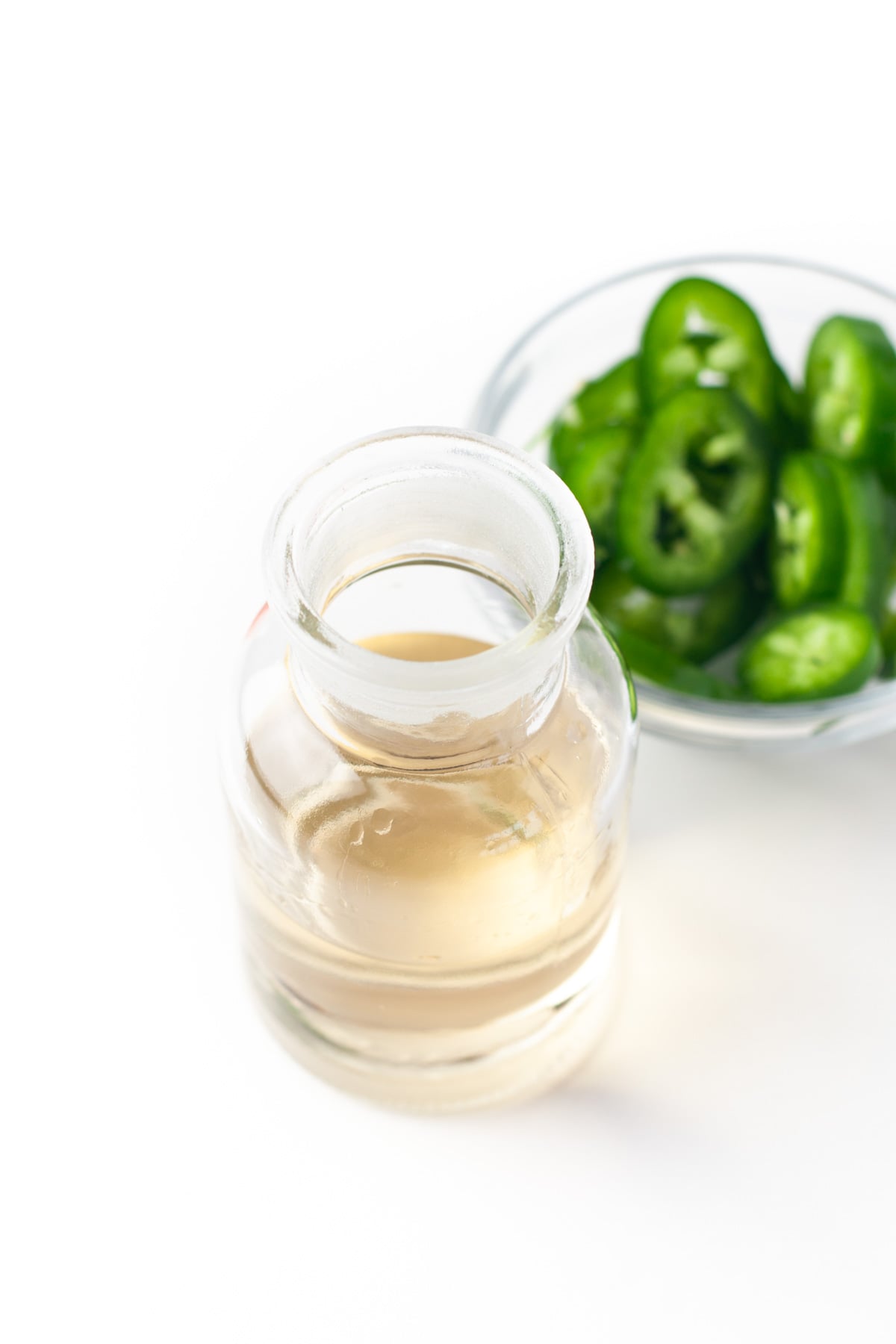 A small glass bottle of clear liquid next to a bowl of sliced green jalape&ntilde;o peppers on a white surface.
