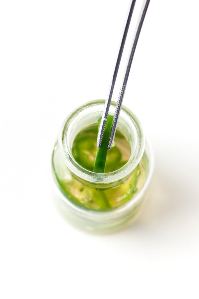 A pair of metal tongs holds a green pickle slice above an open glass jar filled with pickle brine on a white background.