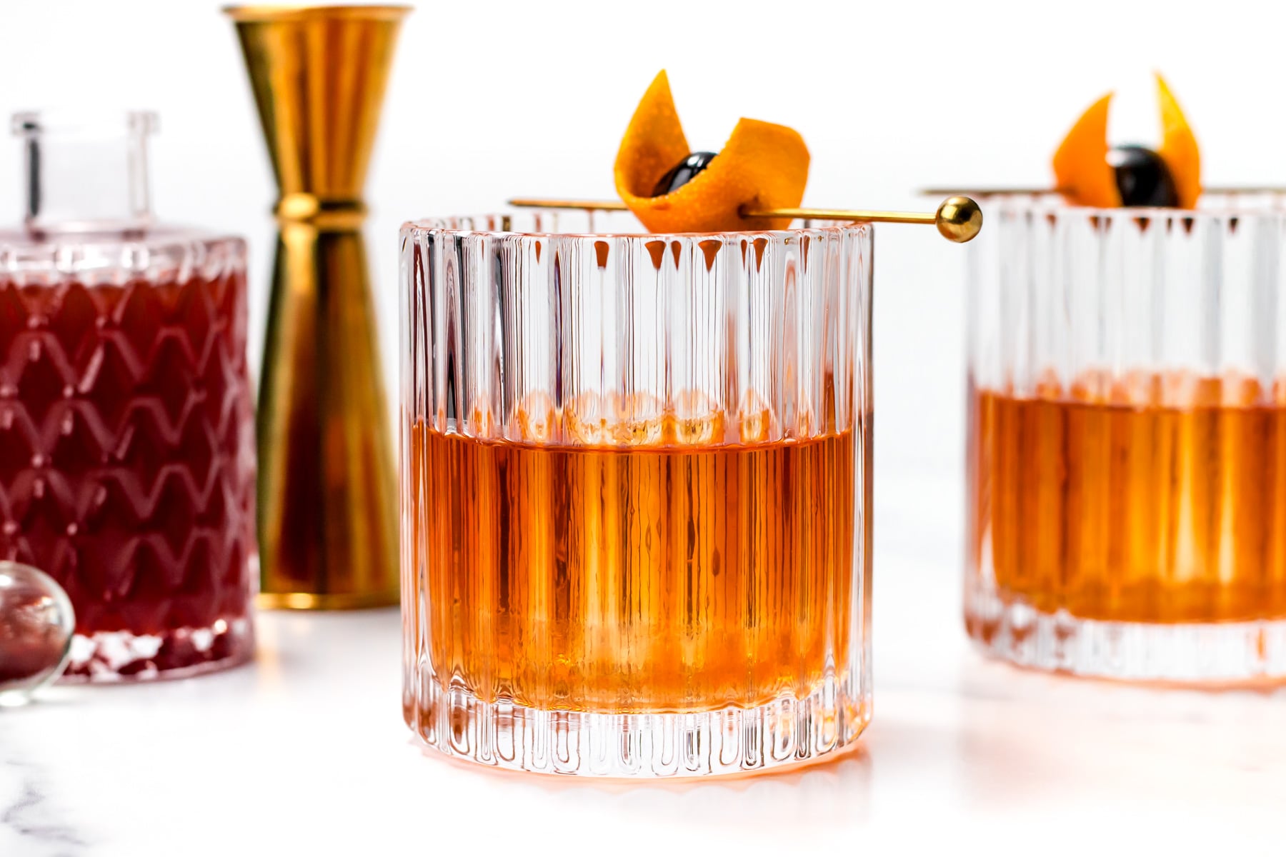 Two old fashioned cocktails in textured glasses, garnished with orange peel and cherries, sit next to a jigger and a decanter on a white surface.