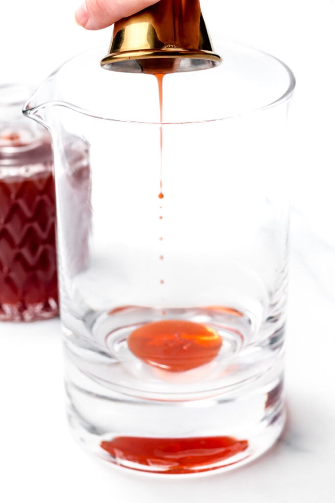 A hand pours red liquid from a gold jigger into a large clear glass mixing pitcher. A glass container with more liquid is in the background.