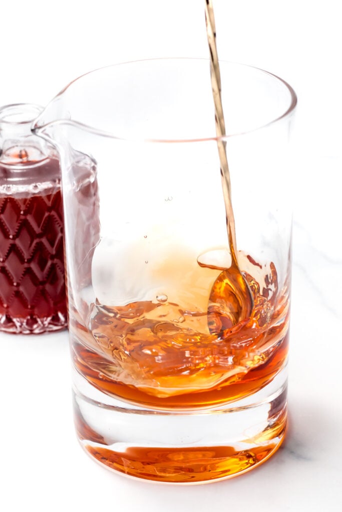 Liquid being poured into a glass mixing vessel containing amber-colored liquid, with a small bottle of red liquid in the background.