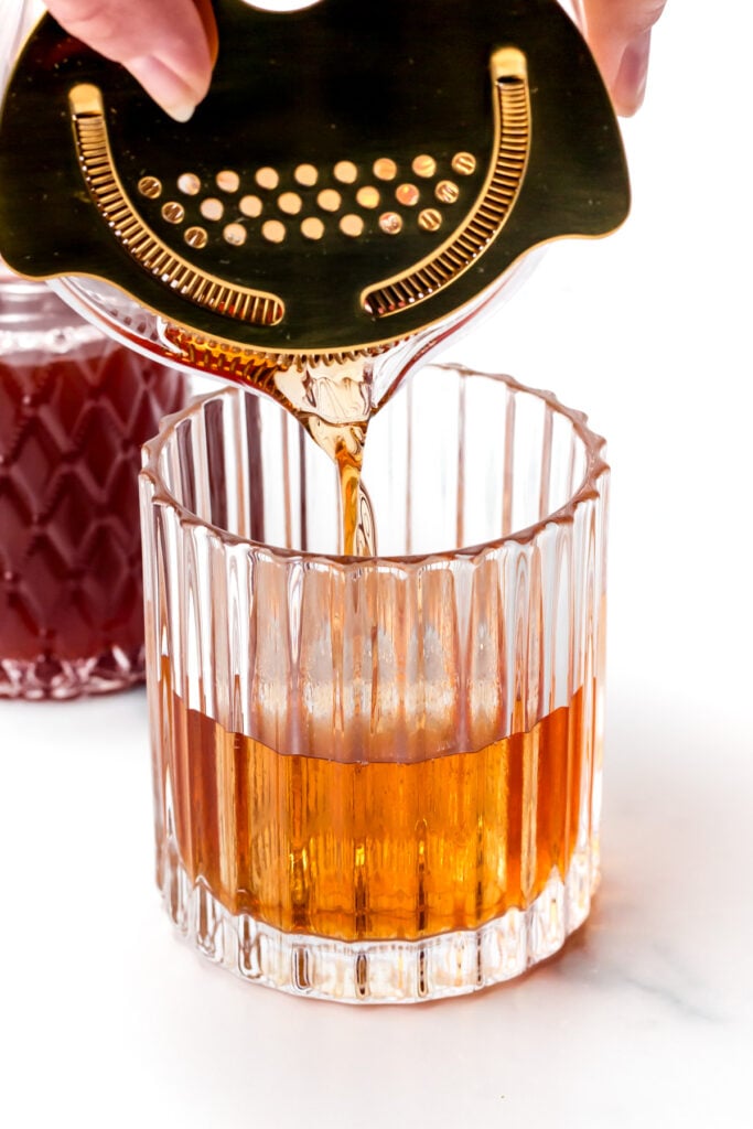 A hand pours a golden-brown cocktail through a strainer into a clear glass with vertical ridges; a bottle with a dark liquid is in the background.