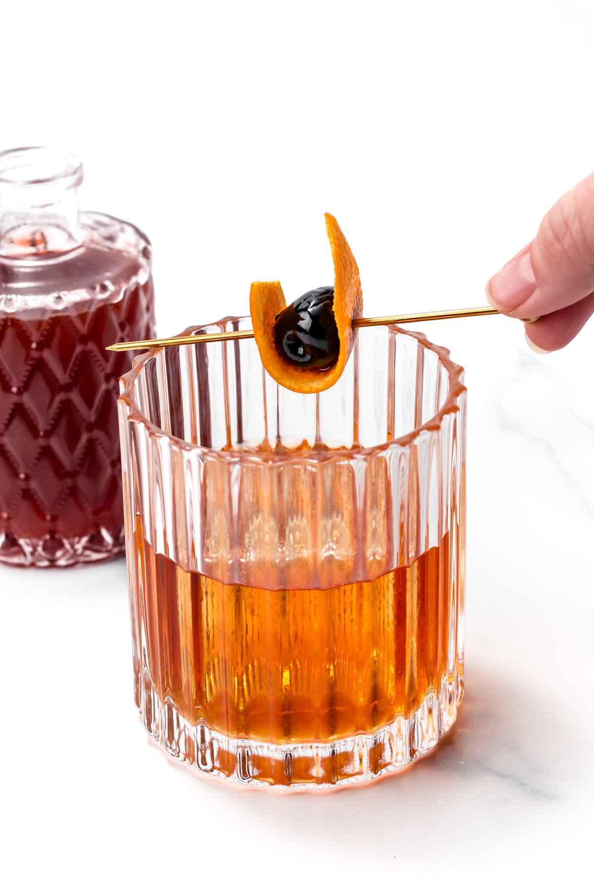 A hand holds a cocktail pick with an orange twist and cherry over a glass of amber-colored drink; a bottle of dark liquid sits in the background.