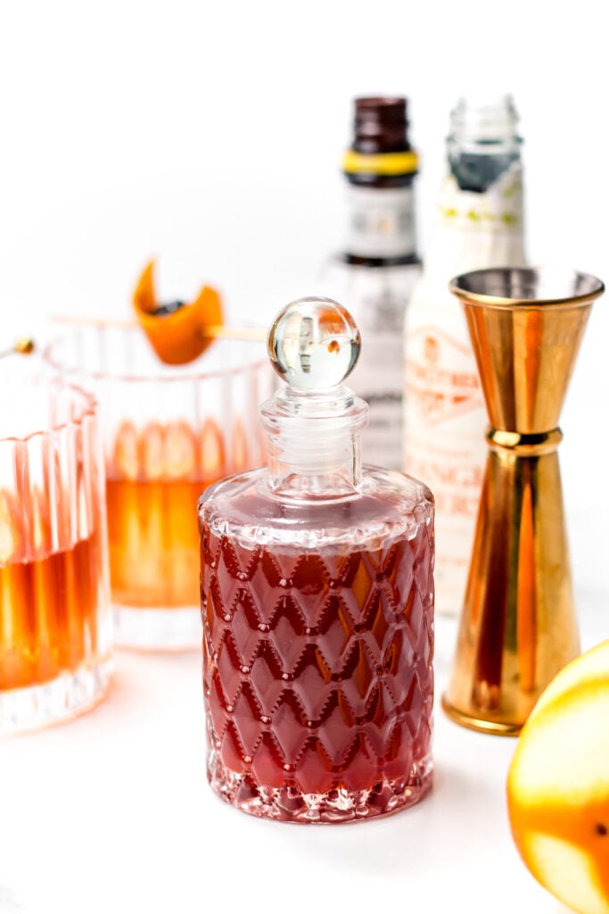 A glass decanter filled with red liquid is surrounded by two cocktail glasses, a jigger, bitters bottles, and an orange garnish on a white surface.