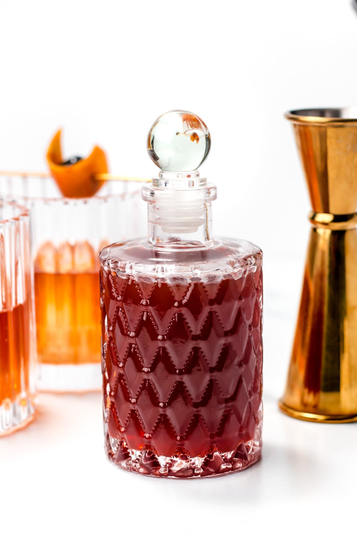 A textured glass decanter filled with a dark red liquid, surrounded by a cocktail, a glass, and a gold jigger on a white background.