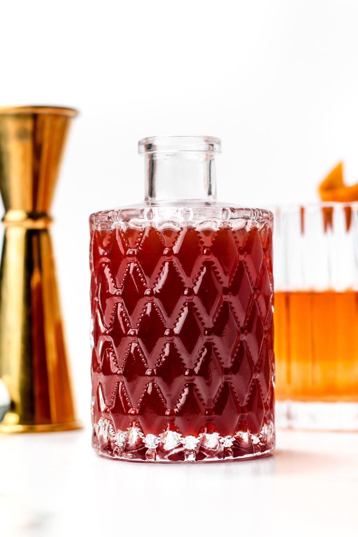 A glass decanter filled with a dark red liquid, placed on a white surface, with a gold jigger and an amber drink in the background.