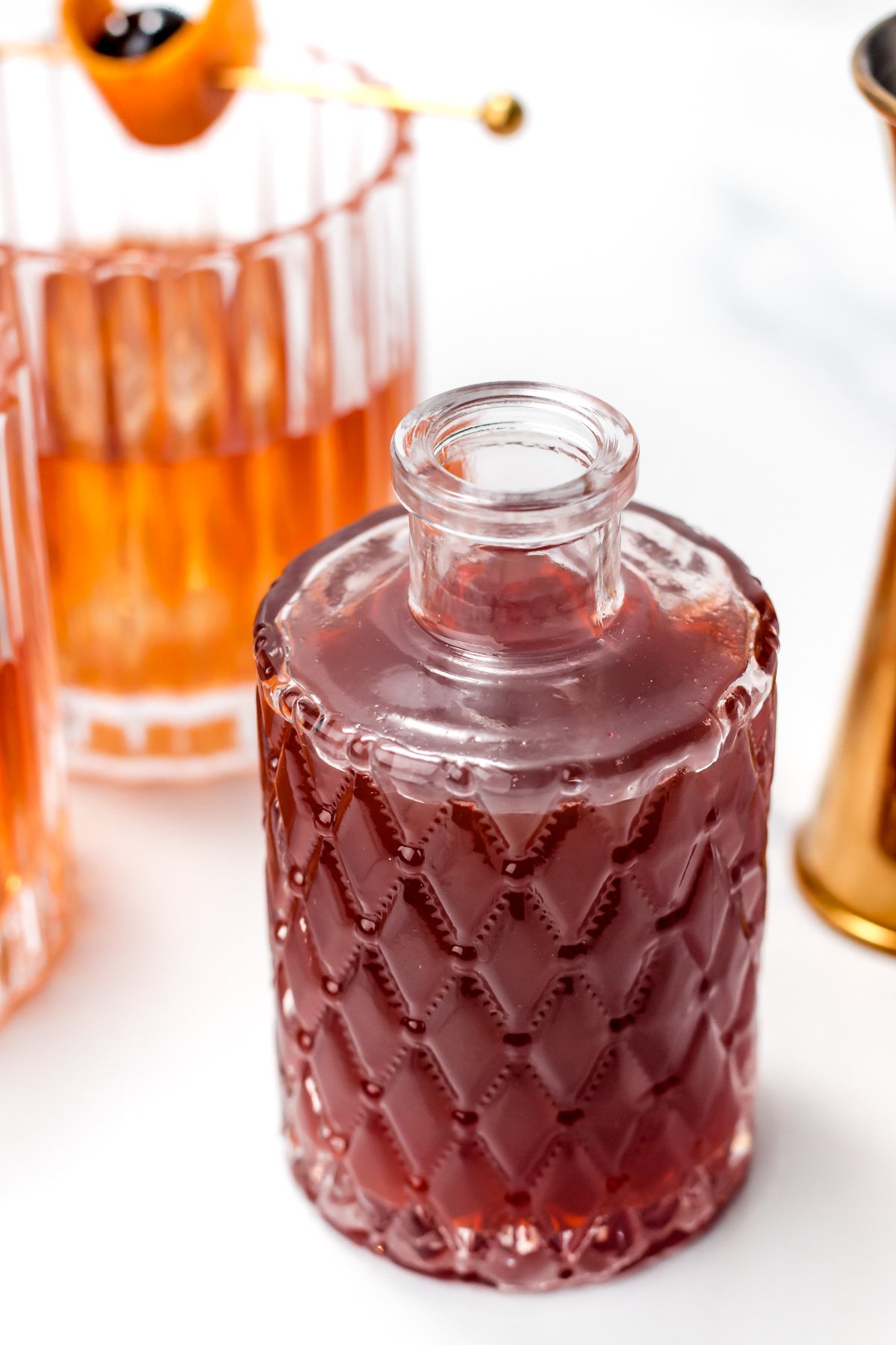 A glass bottle with a diamond-patterned texture containing a dark red liquid, placed next to a cocktail glass and a gold jigger on a white surface.