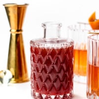 A patterned glass decanter filled with a dark red liquid is placed between a gold jigger and two cocktails in clear glasses with orange garnishes.