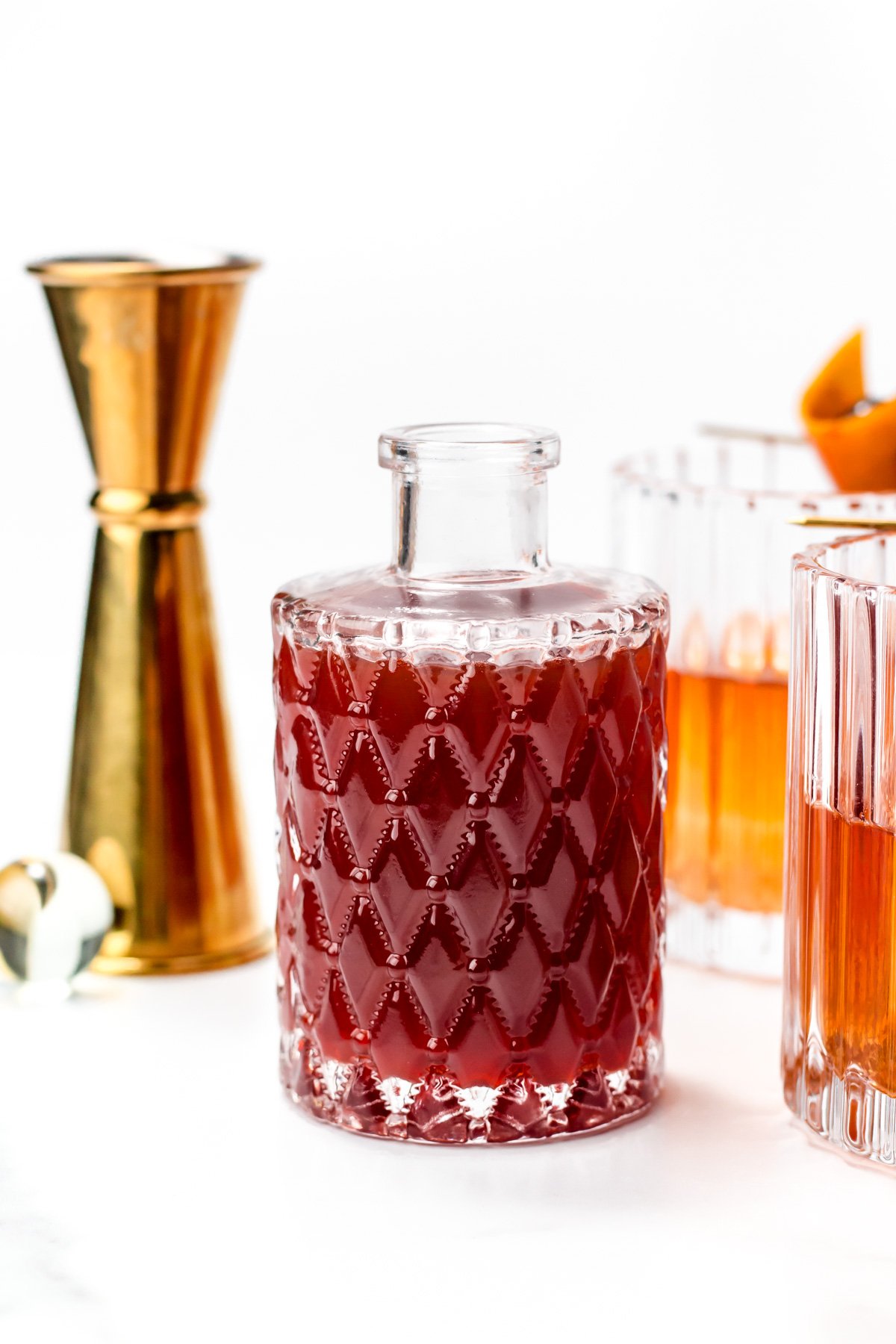 A patterned glass decanter filled with a dark red liquid is placed between a gold jigger and two cocktails in clear glasses with orange garnishes.