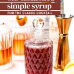A bottle of dark red simple syrup with a glass stopper sits on a white surface, surrounded by cocktail glasses, a jigger, and bottles.