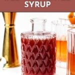 A glass bottle of dark red old-fashioned syrup sits in front of cocktail glasses and a jigger, with text promoting a recipe on feastandwest.com.