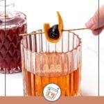 A hand garnishes an old-fashioned cocktail with an orange peel and cherry on a skewer; a bottle of amber liquid is in the background.