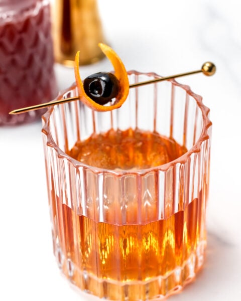 A glass of amber cocktail garnished with an orange peel and a cherry on a gold pick, set on a white surface.