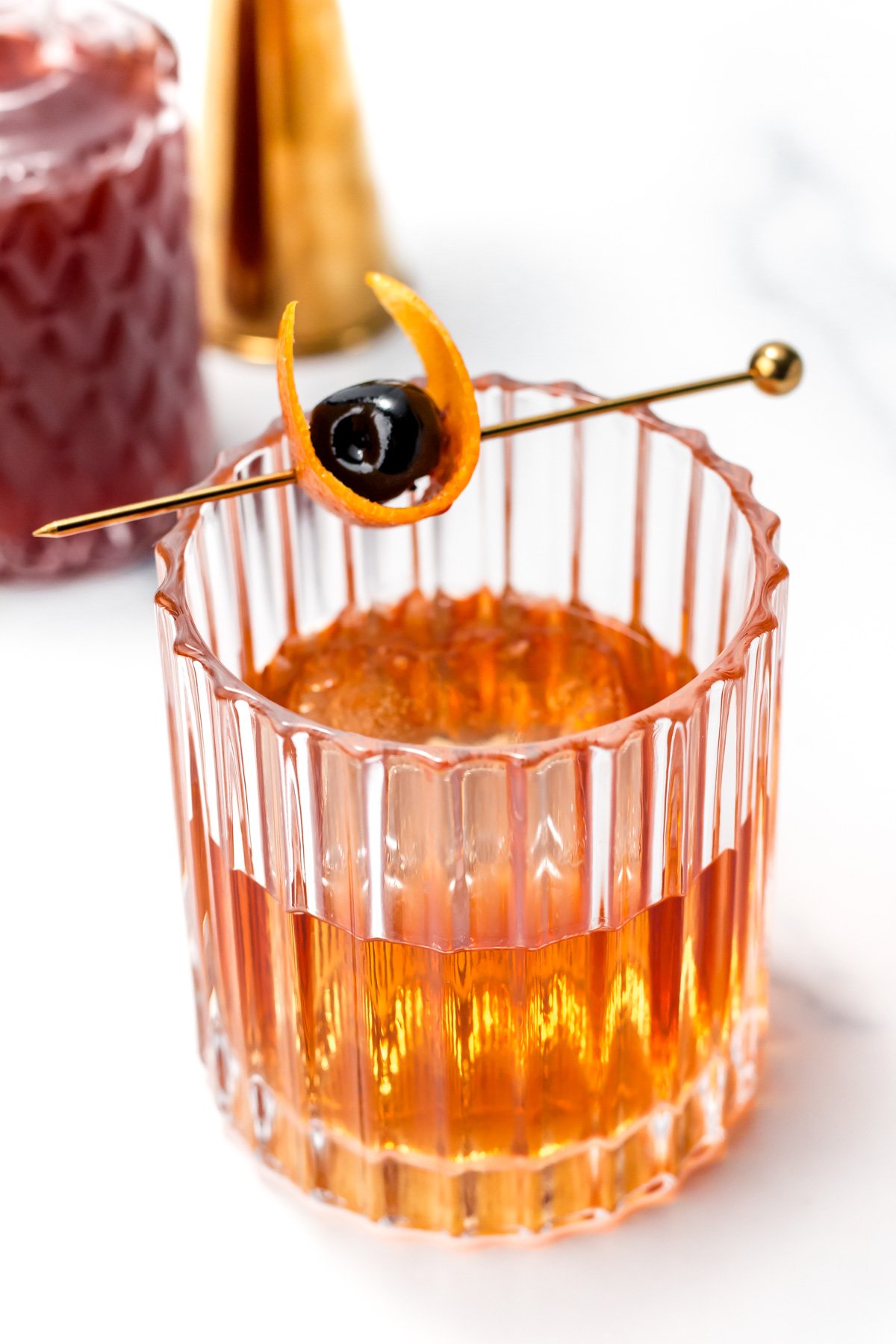 A glass of amber cocktail garnished with an orange peel and a cherry on a gold pick, set on a white surface.