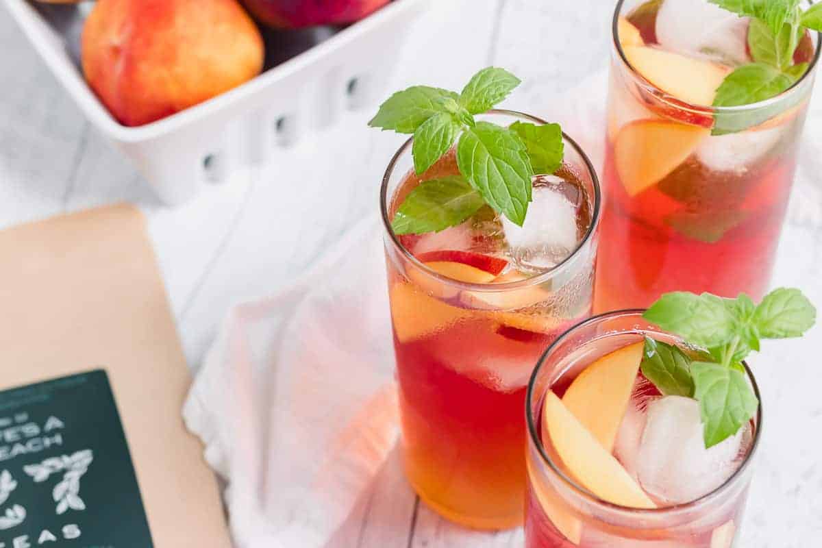 Three glasses of iced peach tea with ice cubes, peach slices, and mint leaves, next to a container of peaches and a package labeled "tea" on a white surface.